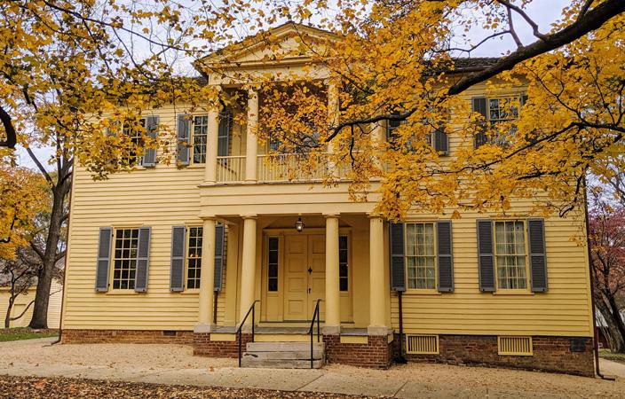 Yellow historic house with blue shutters, framed by autumn trees with golden leaves.