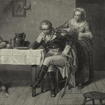 A man sits at a table looking pensive while a woman stands behind him; a dog lies on the floor nearby.