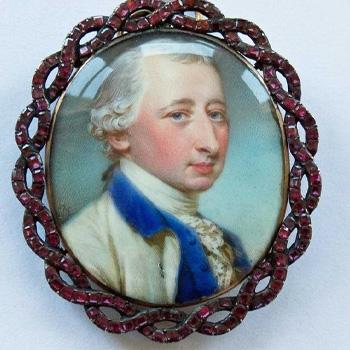 Oval portrait brooch of a man in 18th-century attire, framed with red stones on a decorative border.