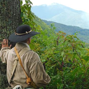 Person in historical clothing and wide-brim hat looks out over a lush, mountainous landscape.