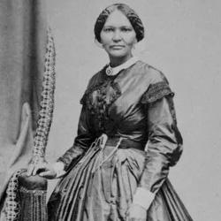 Black and white portrait of a woman in a long dress, standing with one hand resting on a decorative object.