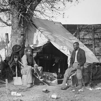 Two men sit outside a canvas tent under a tree, with tools and scattered objects on the ground.
