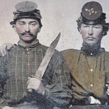 Two young men in old-fashioned uniforms, one holding a large knife, both wearing hats and looking at the camera.