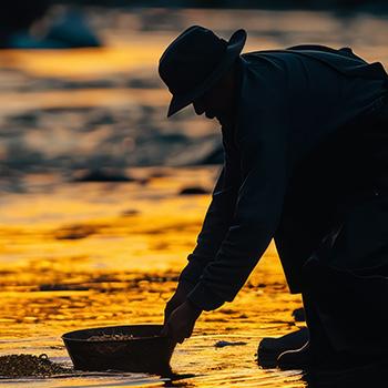 Silhouette of a person panning for gold in a river at sunset with golden light on the water.