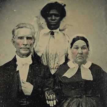 Three people pose for an old portrait: two older white adults sit, with a Black woman standing behind them.